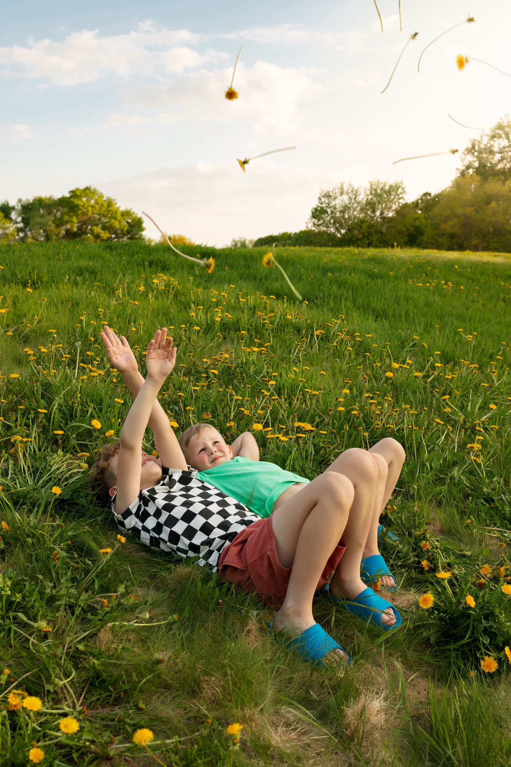 Two young children lying in a grassy field, tossing dandelions.
